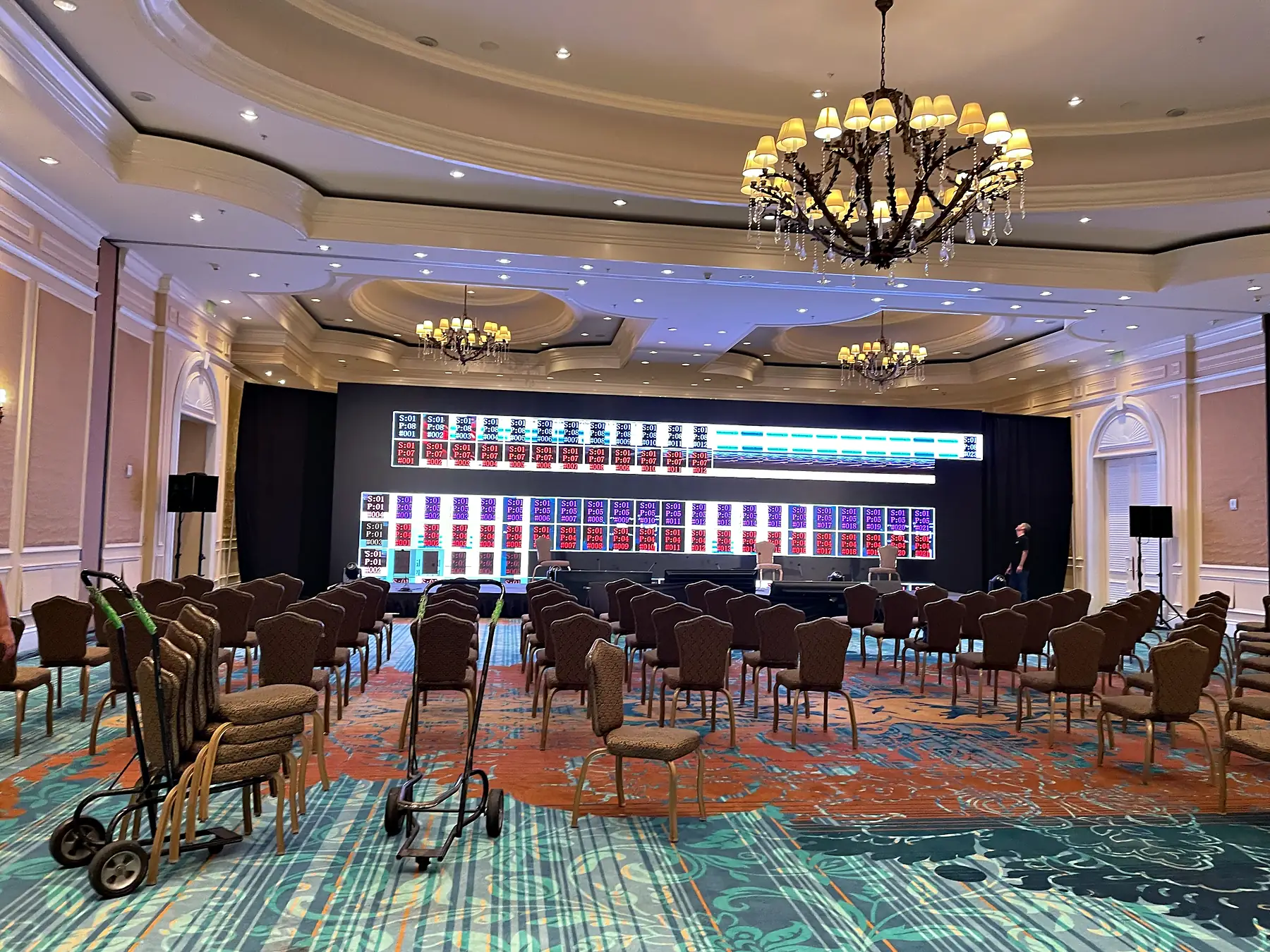 A ballroom under chandeliers with rows of chairs facing a very wide lit LED wall carrying a grid of separate panels, a low stage across the front of it, and loudspeakers on stands at both ends.