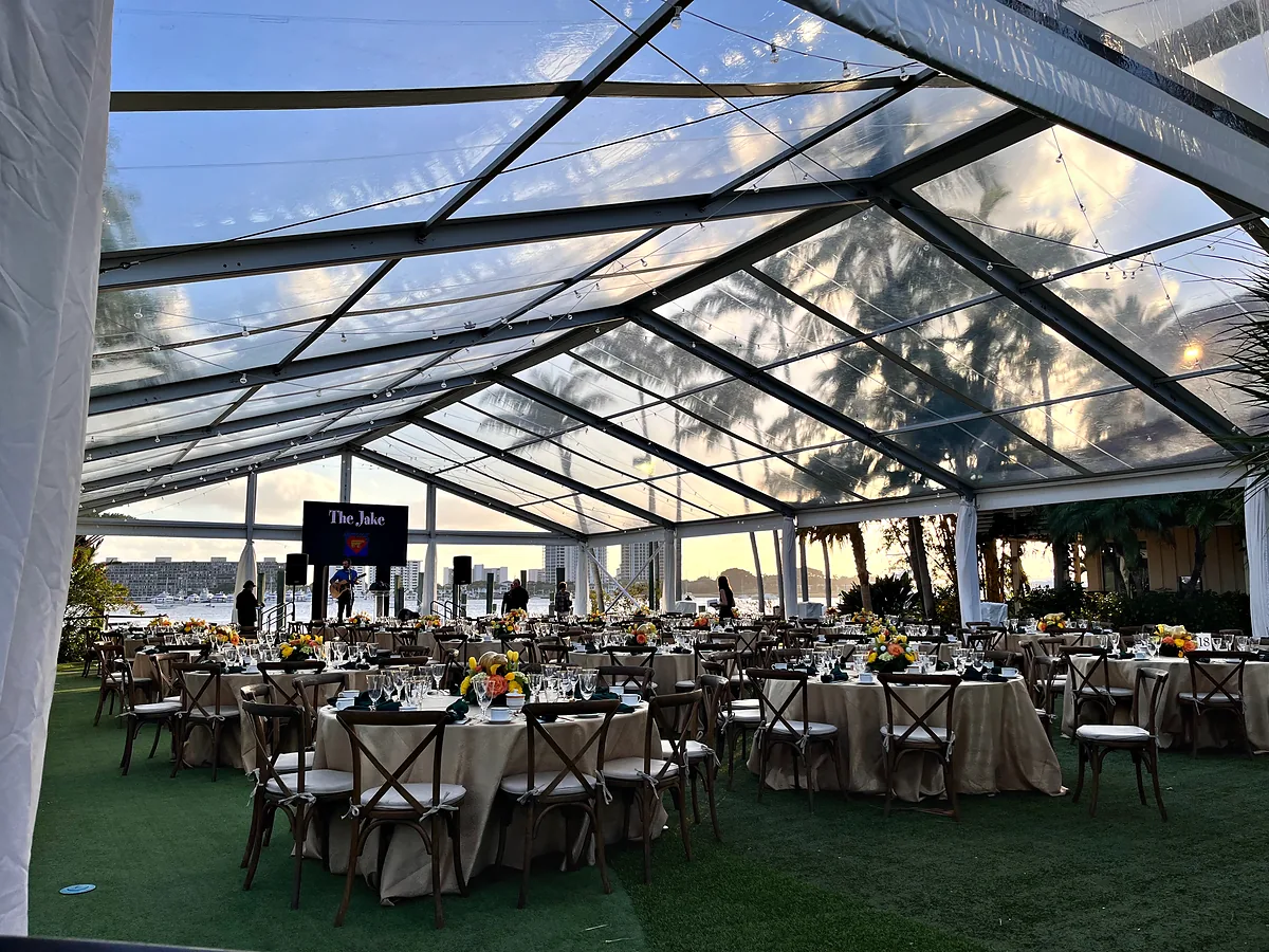 Round tables set on a lawn under a clear-span tent whose roof is open to the sky, a lit display on a stand at the far end, and water and a low shoreline beyond the open side of the tent.