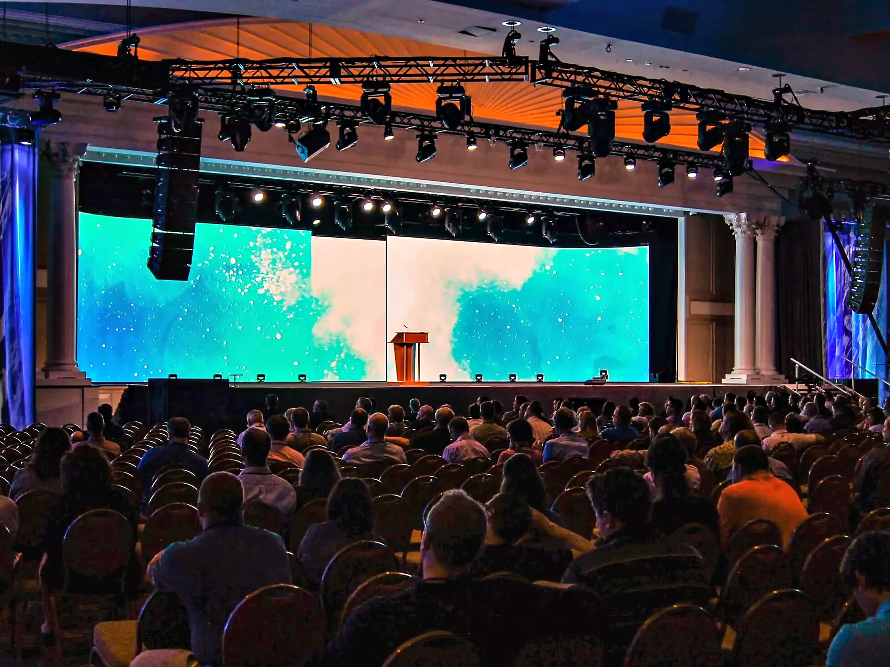 A full house of seated guests facing a stage: a wide video wall carrying a pale blue-green field, a wooden lectern at the center of the deck, a truss of moving lights above it and loudspeakers hung at both sides.