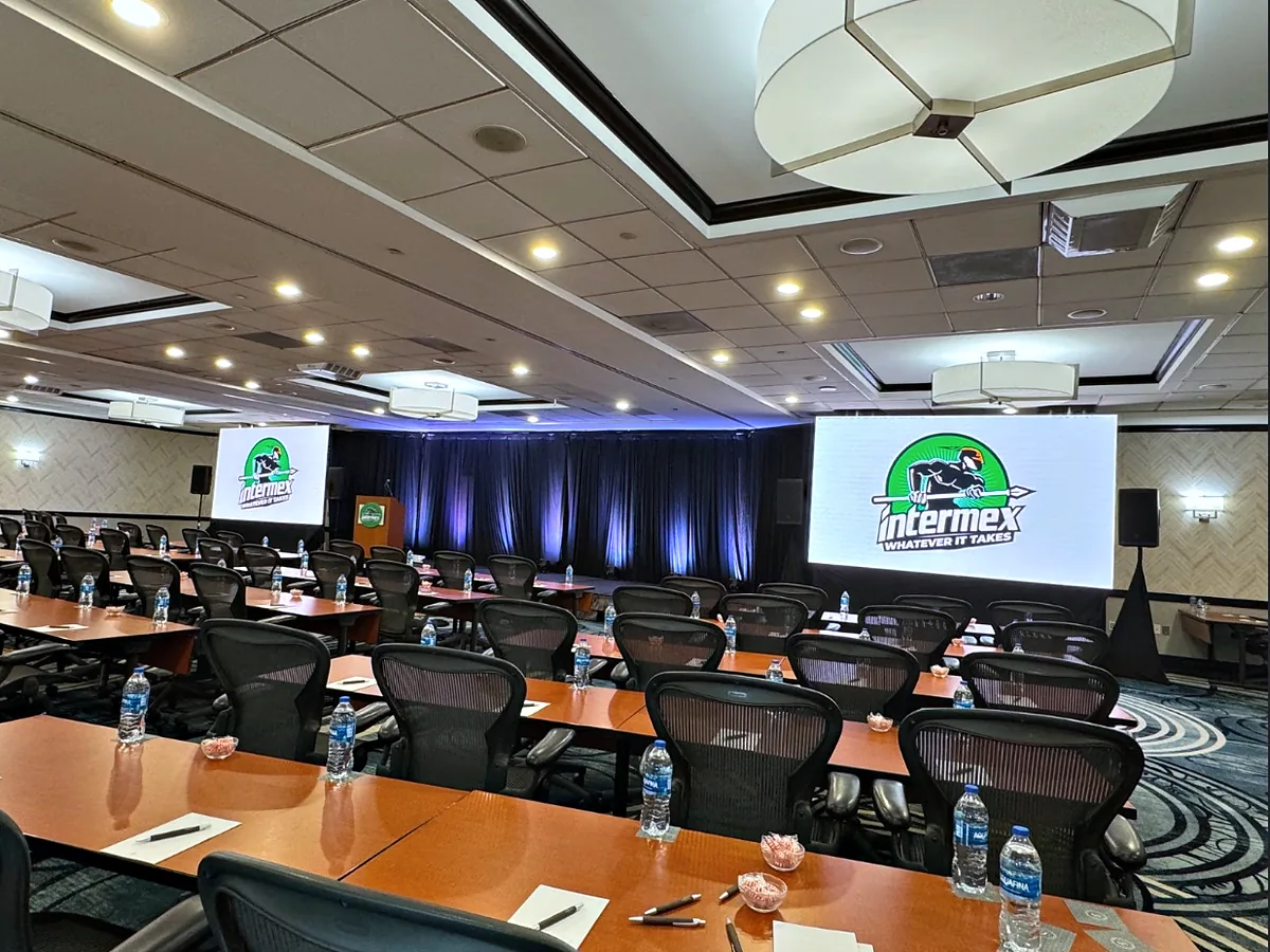 Two video walls carrying the same graphic, one either side of a backlit pipe-and-base drape behind the stage, a lectern in front of the left-hand wall, loudspeakers on stands at both sides, and rows of tables set with water and notepads.