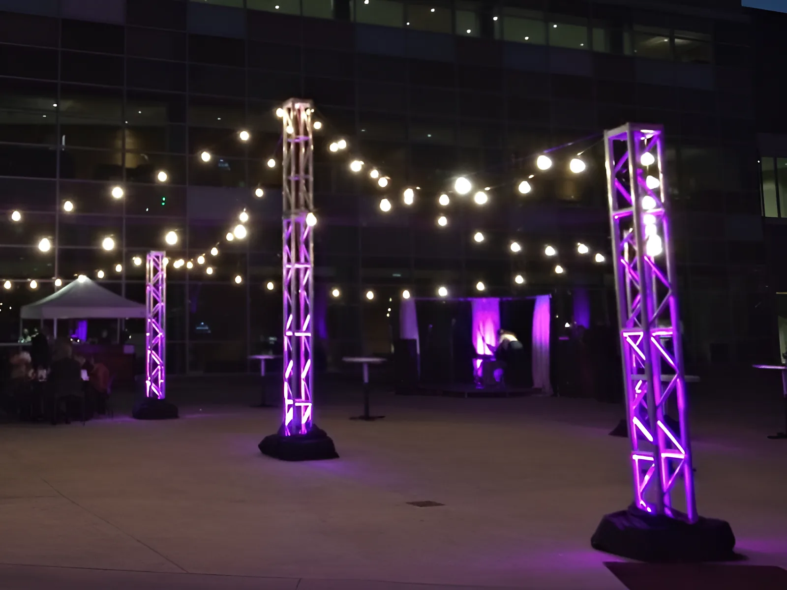 Strings of festoon lighting hung between truss towers washed purple on a terrace at dusk, high tables set out beneath them, and a glass-fronted building behind.