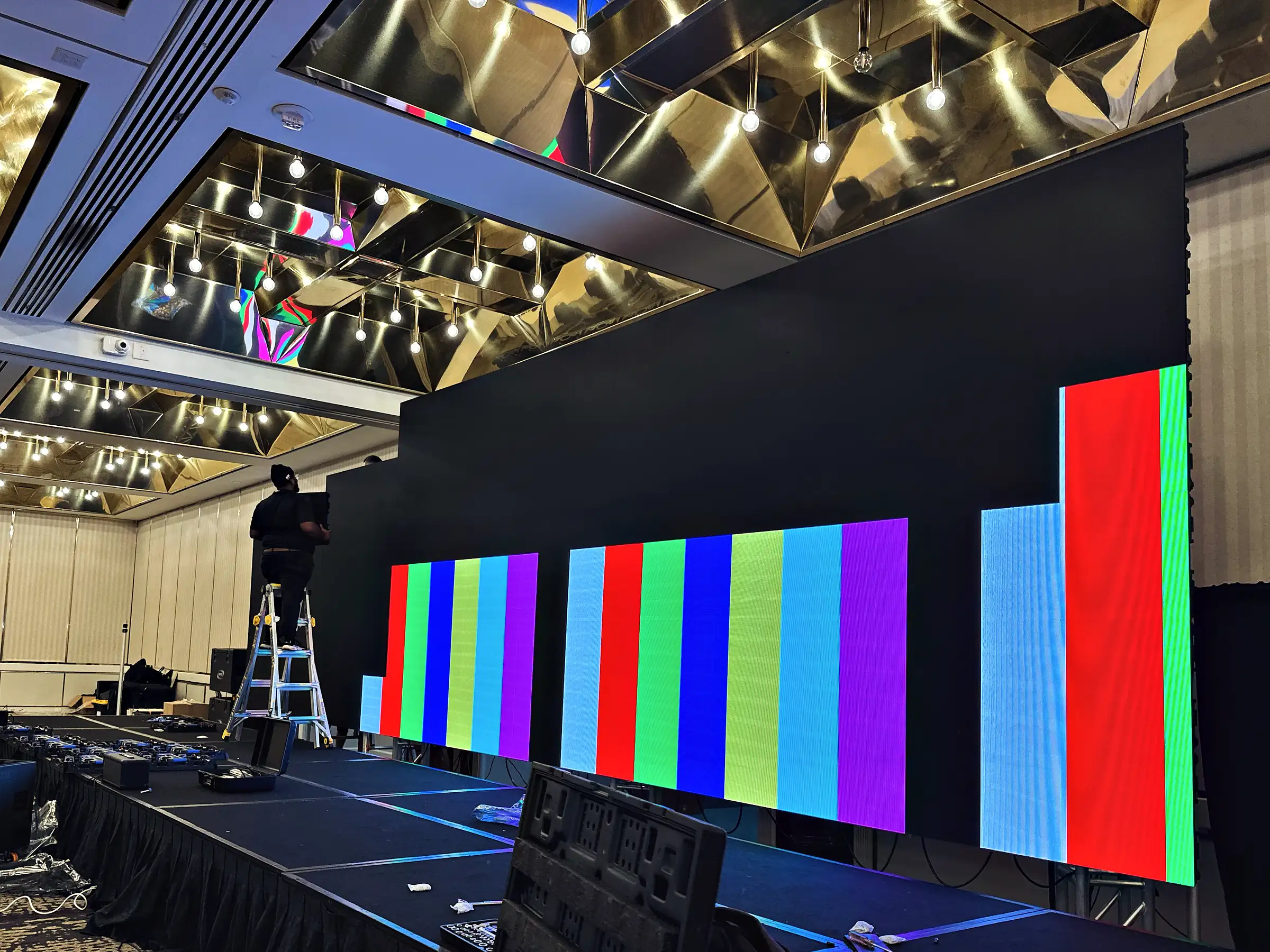 A technician on a step ladder at the end of a long LED video wall built into a black set on a deck, the wall driven to vertical color-bar test fields under a gold faceted ceiling.