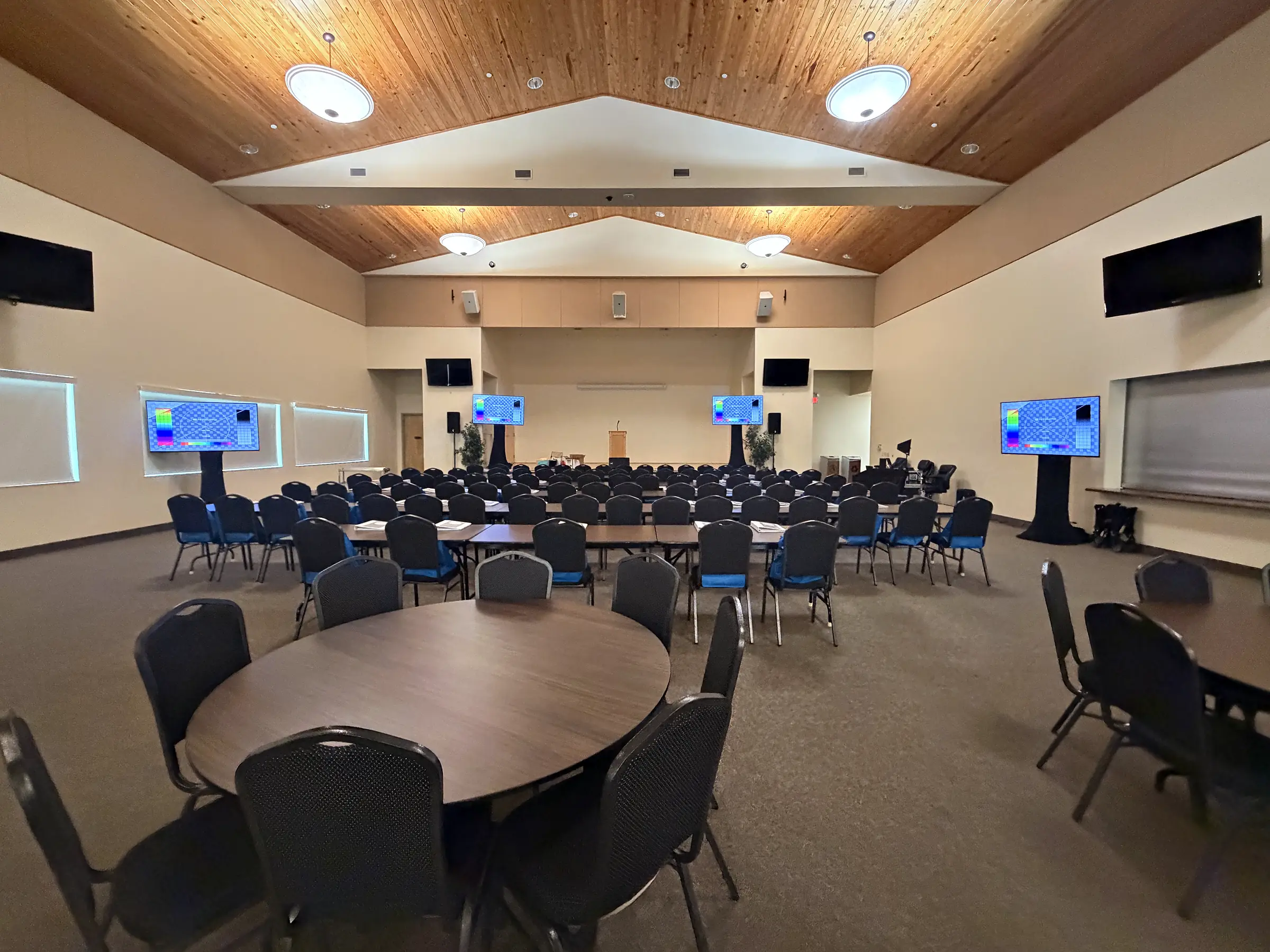 A meeting room under a vaulted wood ceiling: round tables and rows of chairs facing the front, flat-panel displays mounted on the side walls and one standing on a rolling frame at the right, and loudspeakers high on the walls.