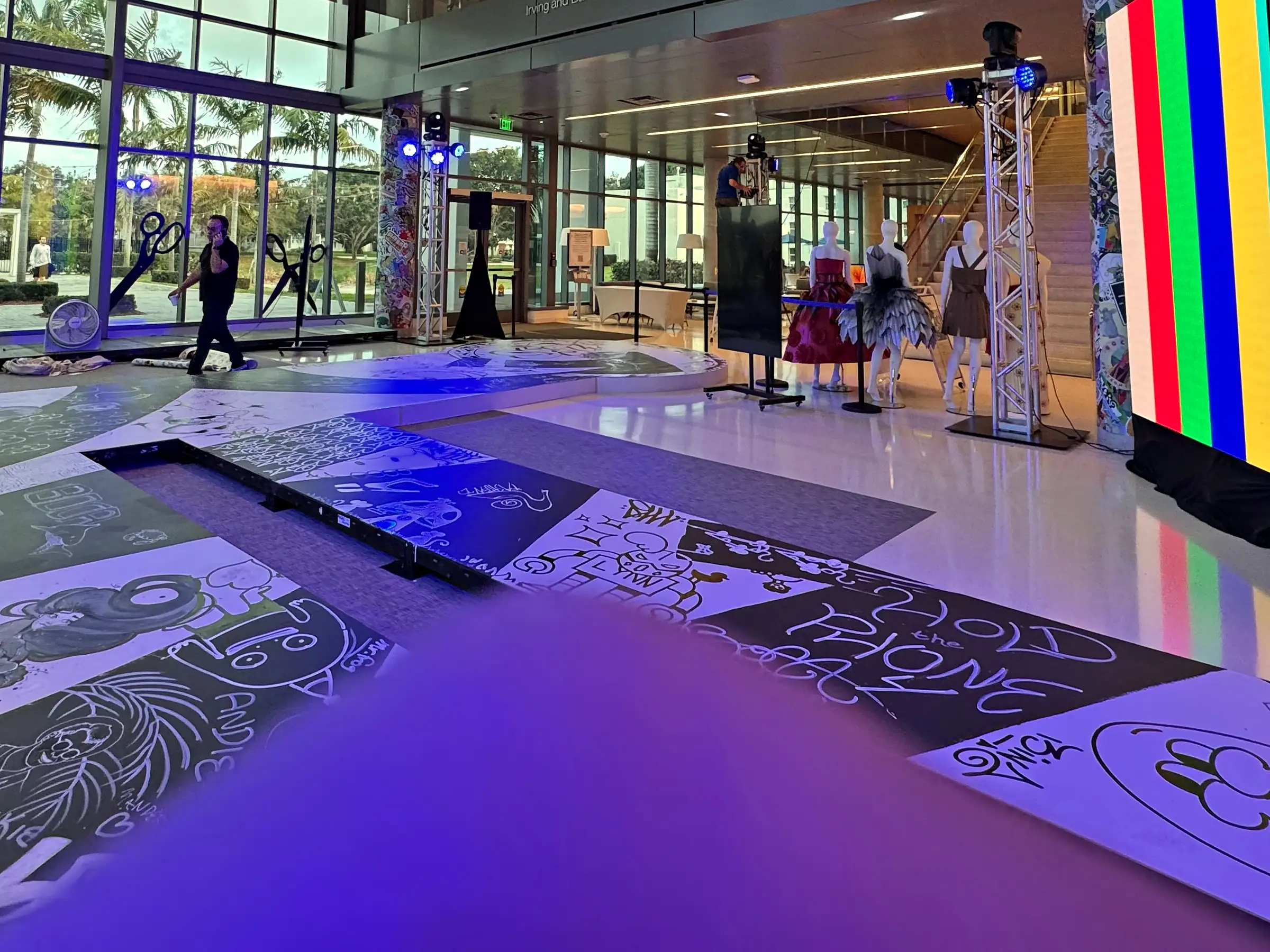 A runway of printed floor panels laid through a glass-walled atrium washed purple, truss towers of moving lights either side, dressed mannequins at the far end, and an LED wall driven to color test fields at the right.