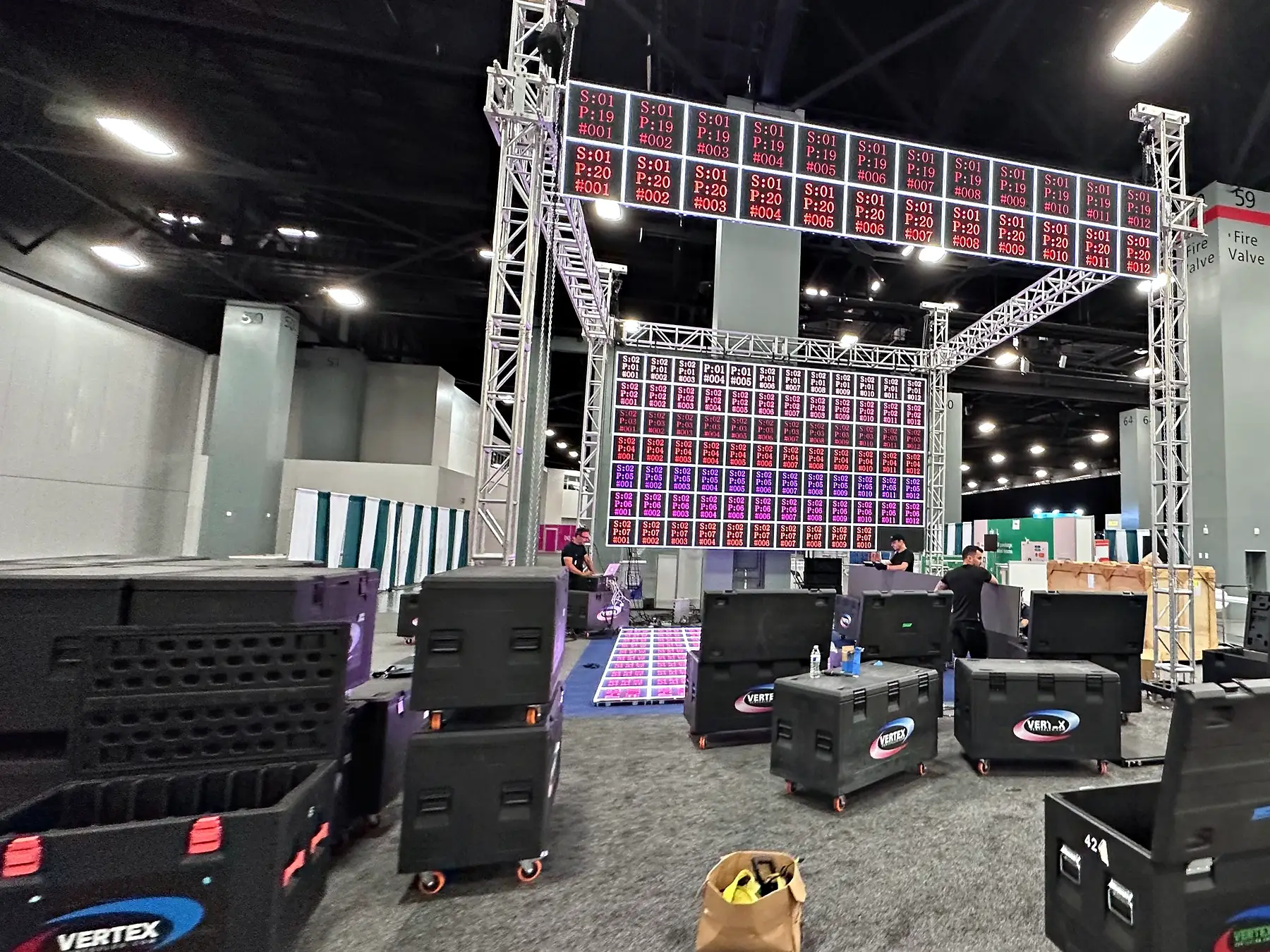 A booth going up on an exhibit floor: two truss towers carrying an overhead header and a back wall of LED panels, all driven to addressing test patterns, road cases across the carpet in front and crew working at the base of the structure.